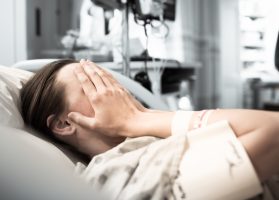 stressed woman holds head while lying in hospital bed
